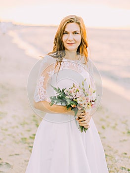 Smiling bride is holding the wedding bouquet of the roses at the background of the sea. The close-up portrait.