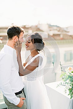 Smiling bride touches groom face with hands on terrace at home