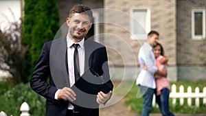 Smiling broker standing with documents, happy family hugging near their new home