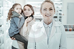 Smiling brown-eyed mature female druggist wearing a white coat feeling joyful