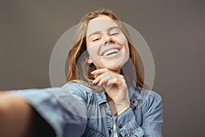 Smiling brown-haired girl dressed in a white t-shirt and jeans shirt makes a selfie on a gray background