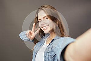 Smiling brown-haired girl dressed in a white t-shirt and jeans shirt shows a v sing and makes a selfie on a gray