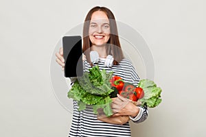 Smiling brown haired woman showing mobile phone with empty display buying products online carries fresh vegetables copy space for