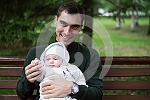 Smiling brunette dad in reen jacket sits on bench with baby in his arms in Park