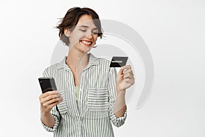 Smiling brunette girl enter credit card info on mobile phone, paying online, making purchase, standing against white