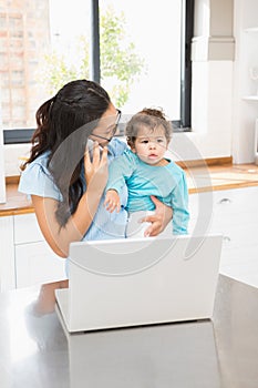 Smiling brunette holding her baby and using laptop on phone call