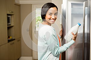 Smiling brunette holding milk bottle with open refrigerator