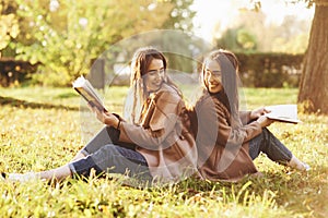 Smiling brunette twin girls sitting back to back on the grass and looking at each other, legs slightly bent in knees