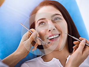 Smiling brunette woman being examined by dentist at sunny dental clinic. Hands of a doctor holding dental instruments