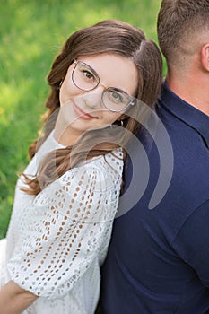 Smiling brunette woman in eyeglasses sitting back to back with unrecognizable man grass in park and looking at camera