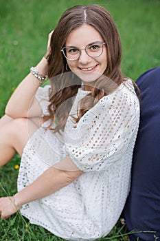 Smiling brunette woman in eyeglasses sitting back to back with unrecognizable man grass in park and looking at camera