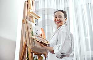 A smiling brunette woman painter holding a palette with paint and a paintbrush and standing in a front of an easel