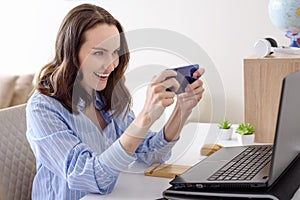 Smiling brunette woman with smartphone in hands sitting at laptop, video games, video calls concept