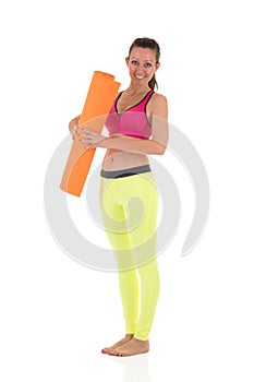 Smiling brunette woman in sports neon yellow leggings and pink bra standing with the orange rolled mat after training