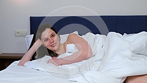 Smiling brunette young woman laying in bed and looking at camera in hotel room.