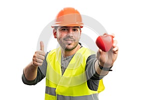 Smiling builder presenting red heart making thumb-up gesture