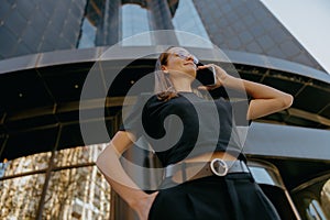 Smiling business woman in eyeglases is talking mobile phone while standing on building background