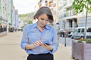 Smiling business woman reading typing looking at smartphone screen walking along city street