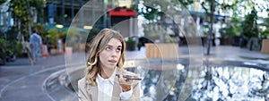 Smiling business woman record voice message, speaking into microphone on mobile phone, sitting near fountain on street