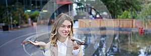 Smiling business woman record voice message, speaking into microphone on mobile phone, sitting near fountain on street