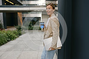 Smiling business woman standing with laptop and take away coffee on stylish office yard