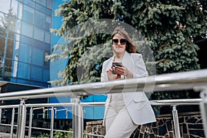 Smiling business woman in white suit and sunglasses using phone during break standing near modern office building