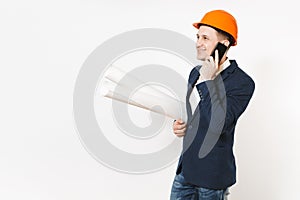 Smiling businessman in dark suit, protective hardhat holding blueprints plans, talking on mobile phone and looking aside