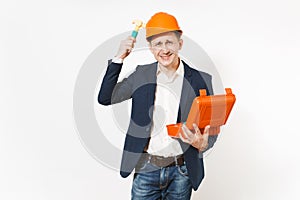 Smiling businessman in dark suit, protective hardhat holding case with instruments or toolbox and beating himself on