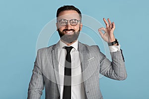 Smiling businessman in formal suit showing okay gesture on blue background