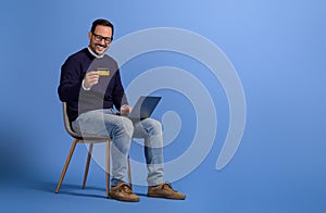 Smiling businessman paying with credit card over laptop computer on chair against blue background