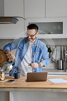 Smiling businessman pouring hot water in coffee cup while working over laptop on kitchen counter at home