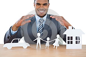 Smiling businessman protecting paper cut out family, house and car with hands