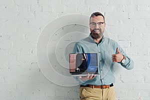 Smiling businessman showing thumb up and holding laptop with booking website