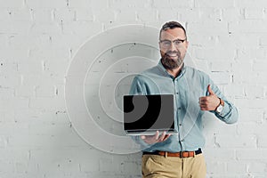 Smiling businessman showing thumb up and holding laptop with copy space