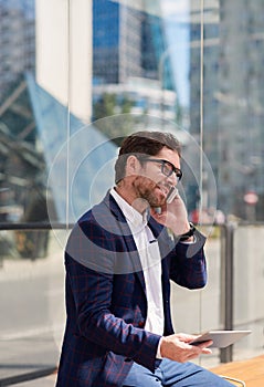 Smiling businessman sitting on a bench using a tablet and cellphone