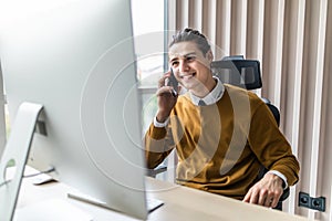 Smiling businessman talking on mobile phone while working on computer. Man sitting at his work table working on computer at home