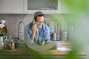 Smiling businessman talking over mobile phone and using laptop on counter in kitchen at home