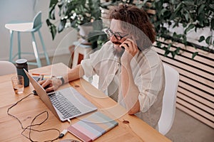 Smiling businessman talking by phone with client while working on laptop in cozy coworking