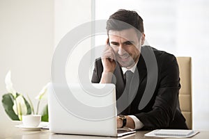 Smiling businessman talking on phone sitting at workplace near l