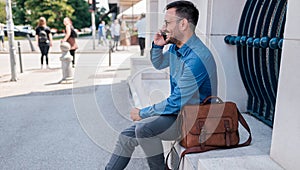 Smiling businessman talking on smart phone while sitting against wall in the city