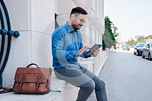 Smiling businessman using digital tablet while sitting against wall on sidewalk