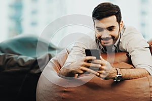 Smiling businessman using and relax smartphone device while sitting on sofa at home.  Man cheerful hipster guy typing an sms messa
