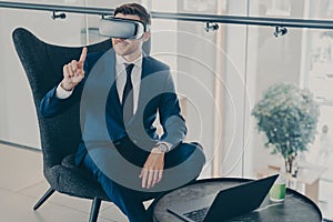 Smiling businessman using VR headset glasses while sitting in office lobby and working on laptop