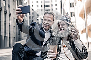 Smiling businessmen in dark costume making selfie with happy homeless