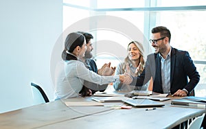 Smiling Businesspeople shaking hands in office with coworkers clapping hands and greeting new employee to their team.