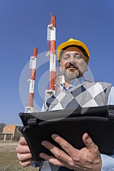 Smiling Businessperson in Yellow Hardhat Holading Digital Tablet and Standing Against Power Plant Chimneys and Blue Sky Background
