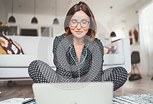 Smiling  Businesswoman dressed pajamas typing notebook keyboard chatting with colleagues sitting cross-legged on living room floor