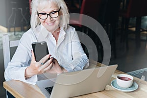 Smiling businesswoman dressed in white shirt is sitting at table in front of laptop and using smartphone. Freelancer