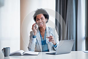 Smiling businesswoman with glass of water talking on smart phone at the office desk