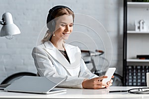 Smiling businesswoman in headphones and formal wear sitting at desk and using smartphone